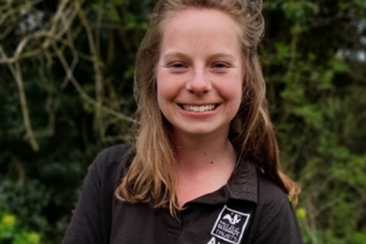 A head and shoulder shot of Esther, who is wearing a black Avon Wildlife Trust polo top. She's stood outside, smiling towards the camera with some trees and greenery behind her.