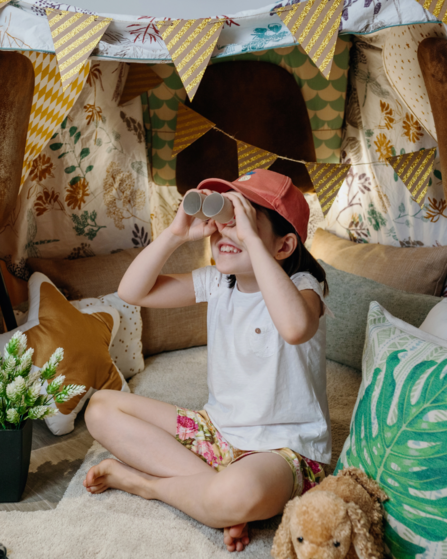 A child sat down, cross-legged in a den, using binoculars made out of cardboard