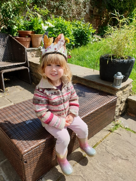 A toddler with blonde hair sits in the garden, wearing a crown made of leaves, feathers and flowers