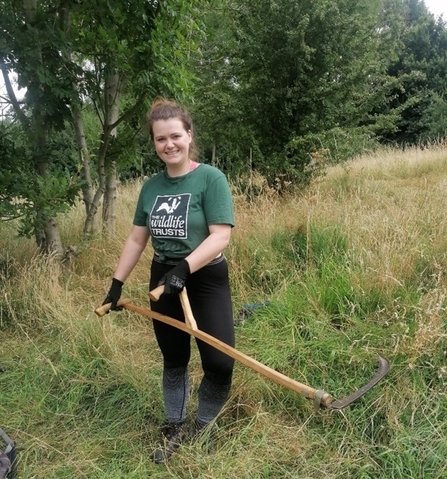 Rebecca is wearing an Avon Wildlife Trust t-shirt, holding a scythe, standing in a field