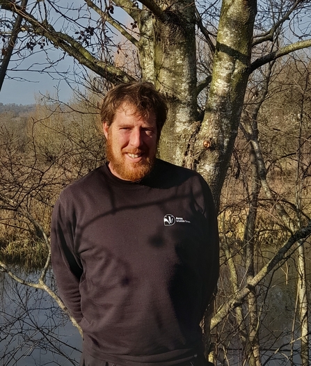 Rob Stephen stood in front of a tree with a river in the background, wearing a black Avon Wildlife Trust sweatshirt