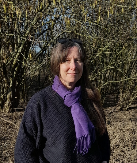 A head and shoulders portrait of Xuela Edwards, standing by a woodland, wearing a purple scarf and black jumper