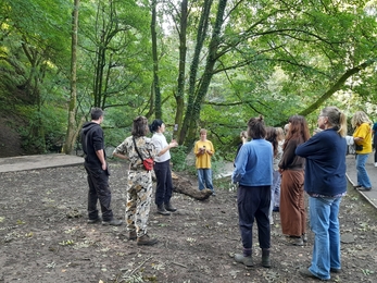 A group of people listening to a woman talk in the woods, all are stood up