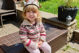 A toddler with blonde hair sits in the garden, wearing a crown made of leaves, feathers and flowers