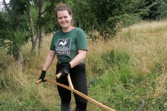 Rebecca is wearing an Avon Wildlife Trust t-shirt, holding a scythe, standing in a field