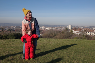 A toddler and her mother stand in a field, with the city in the background. The toddler is pointing across the field.