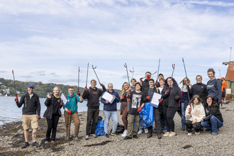 A group photo of young people on a beach clean, they stand on a shingle beach holding litter pickers and bin bags in the air.