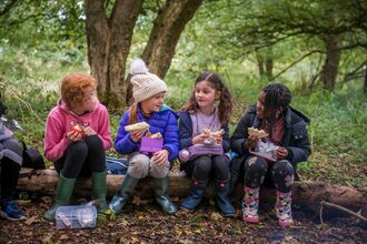 School children sat on a log, smiling and eating a picnic, during education visit to Folly Farm