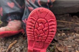 A child is sat on the log in a woodland area. We can see the sole of their wellies, dappled with mud