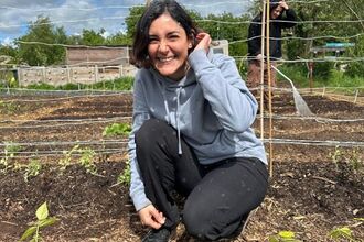 Naz crouches by some food growing fields, smiling at the camera