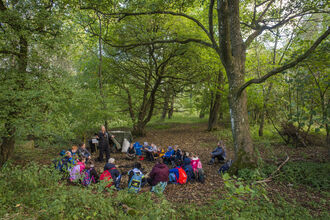 A group of children sat in a circle, in a forest clearing with green trees. Approx 20 children.