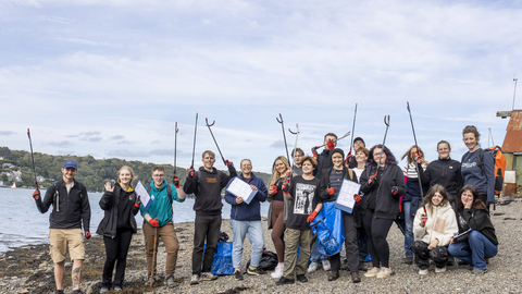 A group photo of young people on a beach clean, they stand on a shingle beach holding litter pickers and bin bags in the air.