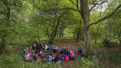 A group of children sat in a circle, in a forest clearing with green trees. Approx 20 children.