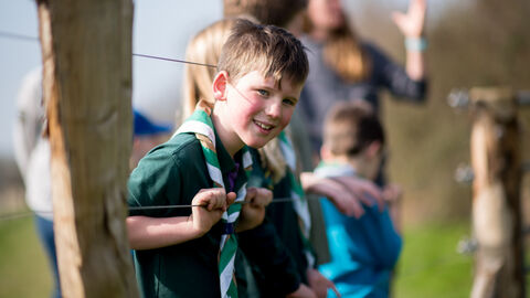 A boy scout smiling at the camera leaning thorugh a fence with wooden posts and wire on a sunny day. People are blurred in the background.