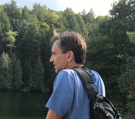 Dominic Hogg wears a blue t-shirt and a black backpack. He is side on, looking towards some trees.