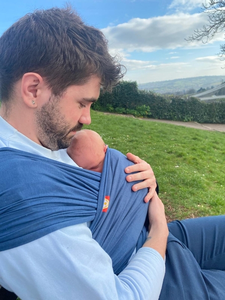 Connor looking down at his newborn daughter in a sling, with a field, trees and rolling hills in the distance.