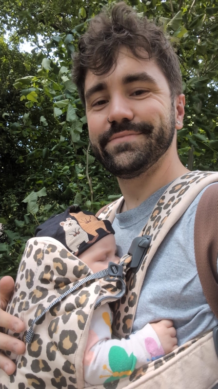 Connor smiles to camera as he wears his newborn daughter in a leopard print carrier.