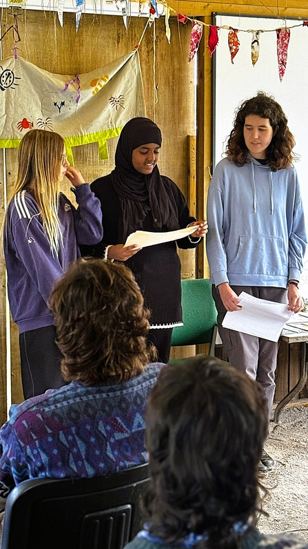 Three young people speaking in front of a group of people with bunting and nature banners around them