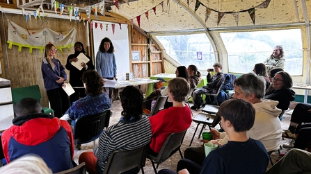 Anature inspired room with three young people speaking in front of a crowd of seated people of all ages, informal, friendly, bunting is on the wall