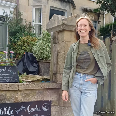 A lady with jeans and a green top and green jacket is smiling, stood next to a wall and fron garden