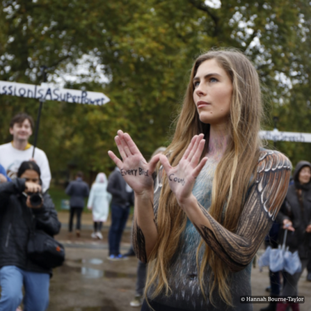A woman with long brown hair with her hands up in front of her chest and body paint on like a bird