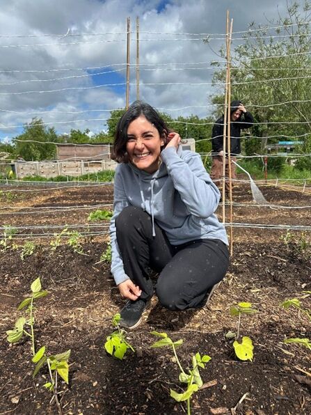 Naz crouches by some food growing fields, smiling at the camera