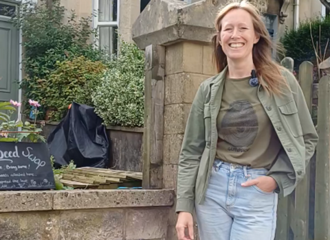 A lady with jeans and a green top and green jacket is smiling, stood next to a wall and fron garden