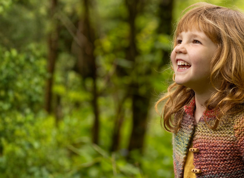 A young girl in a woodland area looks up, smiling in wonder