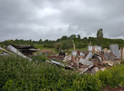 The remains of a demolished building. Piles of rubble and roof material are stacked in the foreground of a green grassland